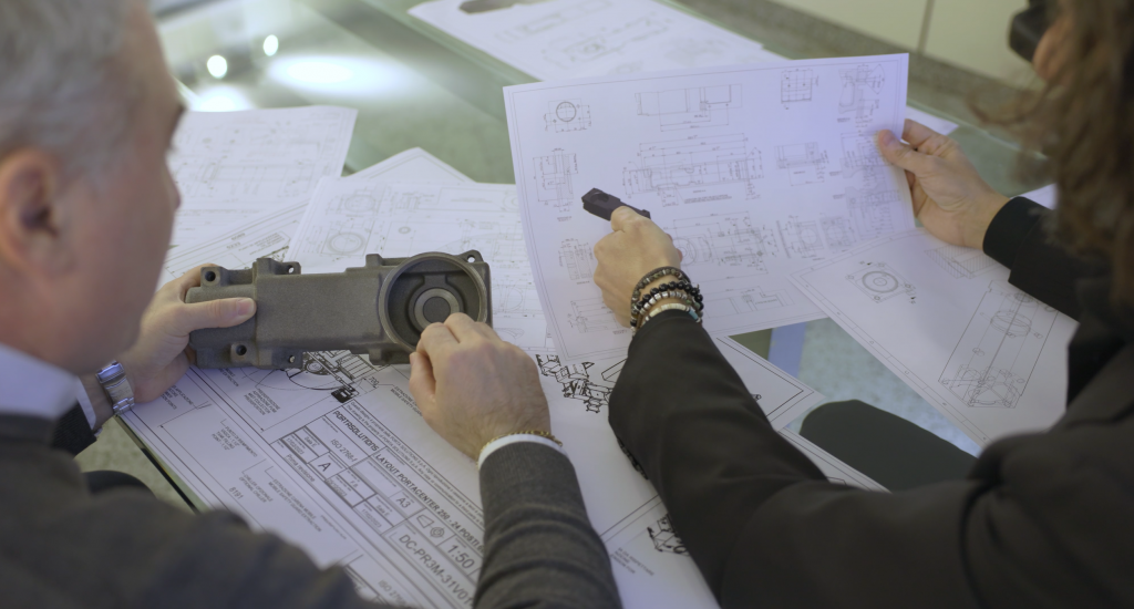Two engineers analyzing a mechanical part and technical drawings on a table